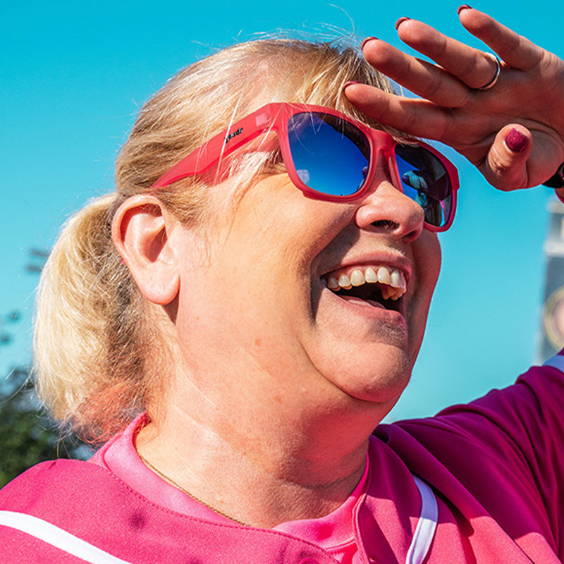 Three-quarter angle view of a lady in a pink baseball uniform & pink sunglasses with blue lenses smiling on a baseball field.