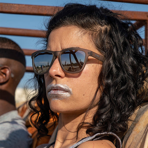 A fierce woman wearing gray sunglasses with gray lenses and silver paint over her mouth drives a rusty post-apocalyptic car.